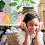 Cheerful Hispanic girl with colored Easter eggs in hands looking at camera while sitting in light room with decorations during Easter celebration